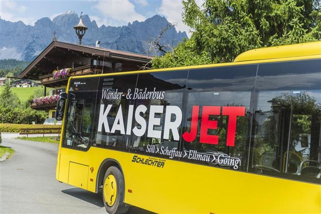 A yellow bus with the inscription "KaiserJET" drives through a picturesque landscape. In the background, mountains and a cozy alpine hut can be seen.