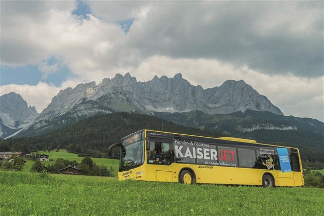 A yellow painted bus is standing on a green meadow, surrounded by impressive mountains. The sky is partly cloudy, creating a picturesque landscape.