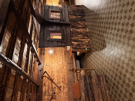 Ein rustikaler Raum mit Holzwänden und einem gefliesten Boden. In der Mitte steht ein Tisch und eine einfache Ausstattung, die eine gemütliche Atmosphäre schafft.