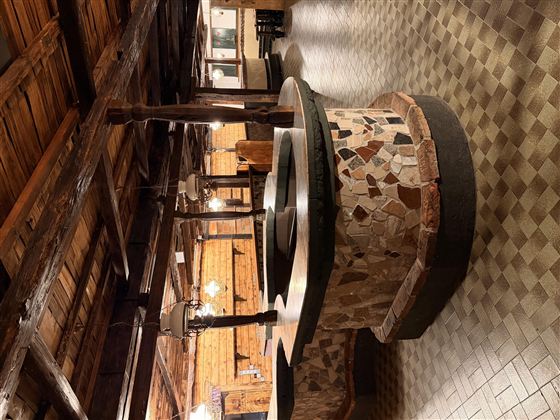 Ein rustikaler Raum mit einem großen, runden Steintisch in der Mitte. Die Wände sind aus Holz und die Böden sind gemustert.