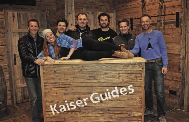 Een groep van zeven personen staat samen aan een houten bar. Ze lachen vriendelijk naar de camera en dragen casual kleding.