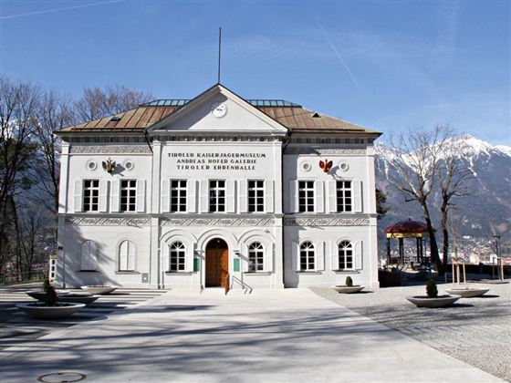 Ein historisches Gebäude mit klassischer Architektur und bunten Verzierungen. Im Hintergrund sind Bäume und ein klarer Himmel zu sehen.