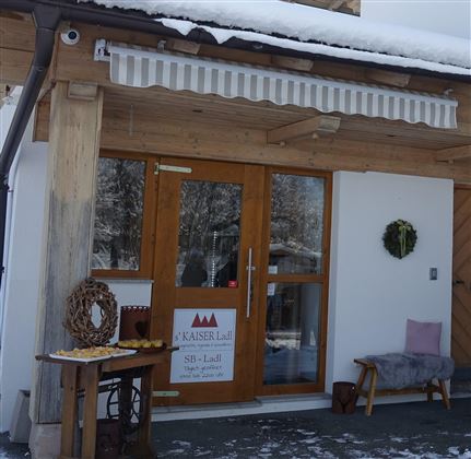 Ein gemütlicher Eingang zu einem Holzhaus mit einem Schild von "Kaiser Lidl". Vor der Tür steht ein Tisch mit Gebäck und eine Sitzgelegenheit.