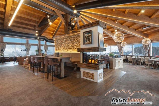 Ein modernes Restaurant mit Holzdekor und großen Fenstern. Die Einrichtung ist gemütlich und einladend, mit einer Bar und einem zentralen Kamin.