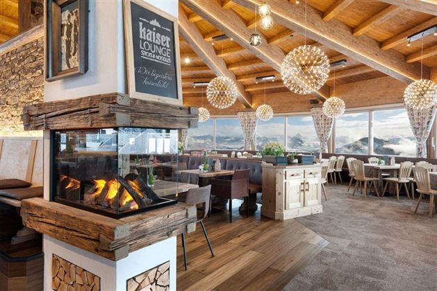 Ein gemütliches Restaurant mit Holzdecke und modernen Lampen. Im Vordergrund befindet sich ein Holzofen, während große Fenster einen Blick auf die Berge bieten.