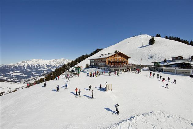 Een besneeuwd landschap met een skigebied en veel skiers. Op de achtergrond zijn bergen en een heldere blauwe lucht te zien.