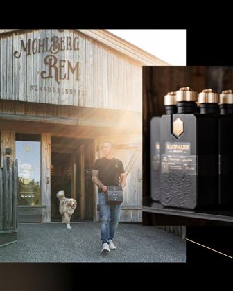 Ein Mann trägt eine Box mit Produkten vor einem Holzgebäude. Ein Hund läuft neben ihm, während die Sonne scheint.