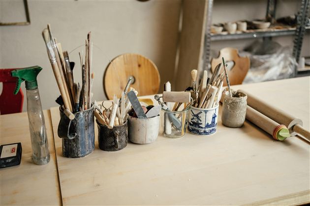 Eine Werkstatt mit verschiedenen Werkzeugen und Pinsel in Behältern. Der Tisch ist aus leichtem Holz und im Hintergrund sind weitere Materialien sichtbar.