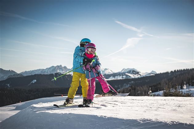 Twee kinderen staan op ski's in de sneeuw. Op de achtergrond zijn besneeuwde bergen en een heldere blauwe lucht te zien.