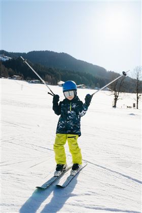 Ein Kind steht auf Skiern im Schnee und hält die Skistöcke hoch. Im Hintergrund sind verschneite Berge und ein blauer Himmel zu sehen.