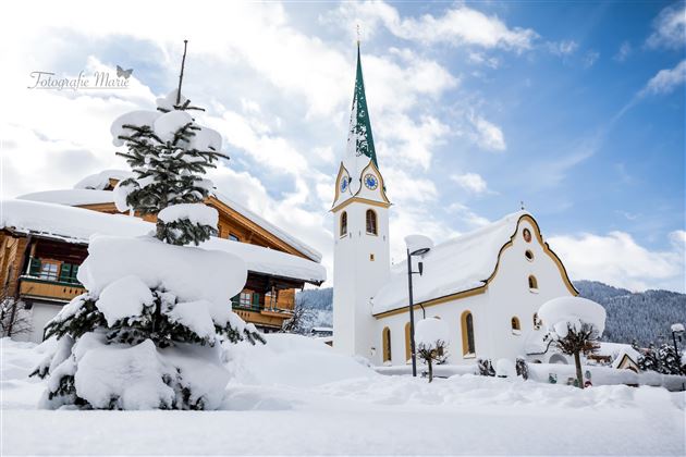 Ein verschneites Dorf mit einer schönen Kirche und einem hohen, grünen Kirchturm. Die Umgebung ist ruhig und winterlich, bedeckt mit frischem Schnee.
