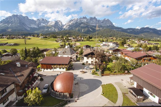 Eine malerische Landschaft mit einem kleinen Dorf im Vordergrund und beeindruckenden Bergen im Hintergrund. Der Himmel ist klar und die Natur ist grün und einladend.