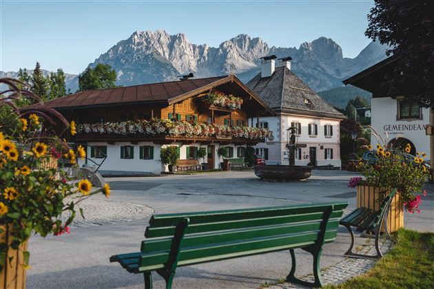 A picturesque village square with traditional wooden houses and blooming flowers. In the background, majestic mountains rise under a clear sky.