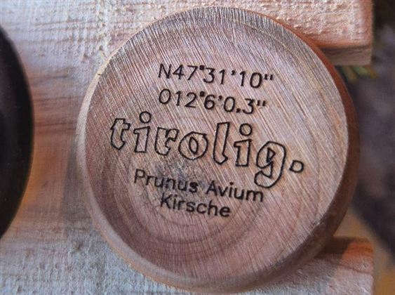 A wooden piece with engraved coordinates and the word "tirolig". Below it is "Prunus Avium" and "Cherry".