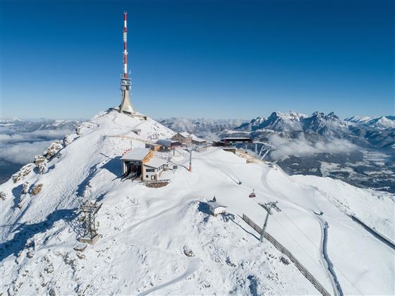 Ein schneebedeckter Berggipfel mit einem Sendemast und einer Berghütte. Im Hintergrund sind majestätische Berge und ein klarer blauer Himmel sichtbar.