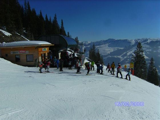 Eine winterliche Landschaft mit viel Schnee und Skifahrern an einer Liftstation. Im Hintergrund sind schneebedeckte Berge und Tannenbäume zu sehen.