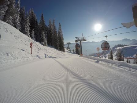 Eine verschneite Piste mit frisch präpariertem Schnee und einem Sessellift im Hintergrund. Die Sonne strahlt über die winterliche Landschaft.