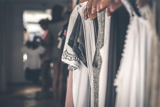 A selection of garments hangs on wooden hangers. Blurred figures are visible in the background.
