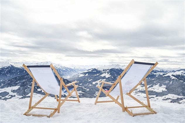Two lounge chairs are on snow-covered ground overlooking snow-capped mountains. The sky is cloudy and the landscape appears calm and peaceful.