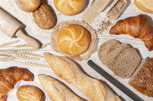 Een verscheidenheid aan vers brood, waaronder croissants en broodjes, ligt op een houten dienblad. Daarnaast zijn tarwekorrels en een deegroller gerangschikt.