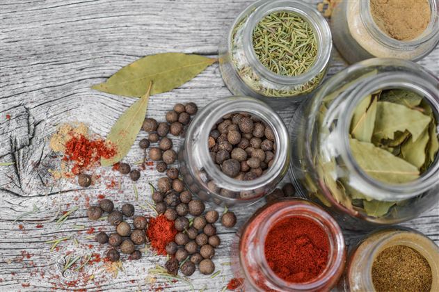 A selection of spices in jars on a wooden table. Various colors and textures are visible, including peppercorns, bay leaves, and paprika.
