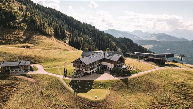 Eine malerische Berghütte umgeben von grünen Wiesen und Wäldern. Im Hintergrund sind majestätische Berge und ein klarer Himmel zu sehen.