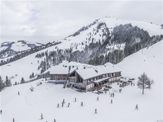 Een pittoresk skigebied met een houten huis en besneeuwde bergen. Veel mensen zijn op de pistes onderweg.
