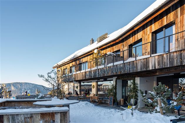 A modern wooden building in the snow with large windows and a clear blue sky. The surroundings are wintry and inviting.