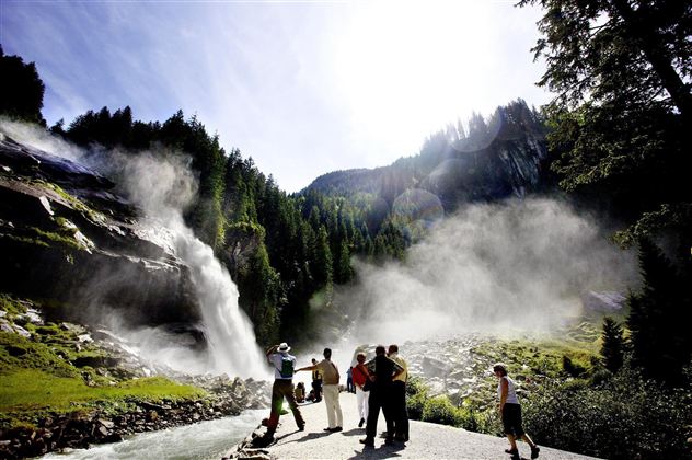 A group of people is standing near an impressive waterfall surrounded by green trees. The sky is clear and the sun is shining brightly.