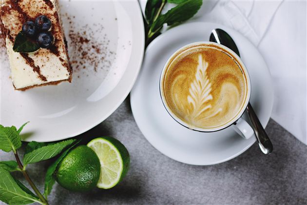 A beautiful plate with a dessert and fresh berries is alongside a cup of coffee with artistic milk foam. Lime and mint leaves complement the arrangement.