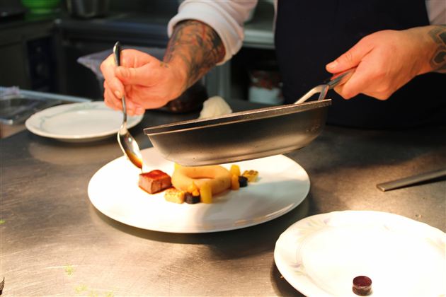 A chef presents an artistic dish on a white plate. Various ingredients are carefully arranged.