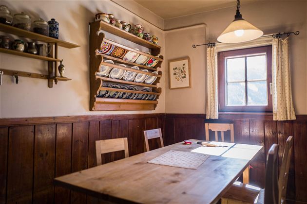 Ein gemütlicher Raum mit einem Holztisch und Stühlen. An der Wand sind Geschirr und Vorratsbehälter zu sehen, während ein Fenster natürliches Licht hereinfluten lässt.