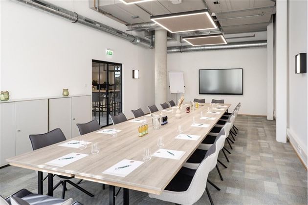 Moderner Konferenzraum mit langem Holztisch, grauen Stühlen, Whiteboard und großem Bildschirm an der Wand.