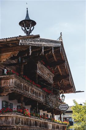 Een traditioneel houten huis met versierde balkons en kleurrijke bloemen. De lucht is helder en de architectuur toont een typische regionale stijl.