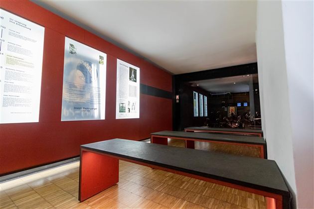 Ein moderner Ausstellungsraum mit roten Wänden und Informationsplakaten. Im Hintergrund sind Sitzbänke und andere Teile des Raumes sichtbar.