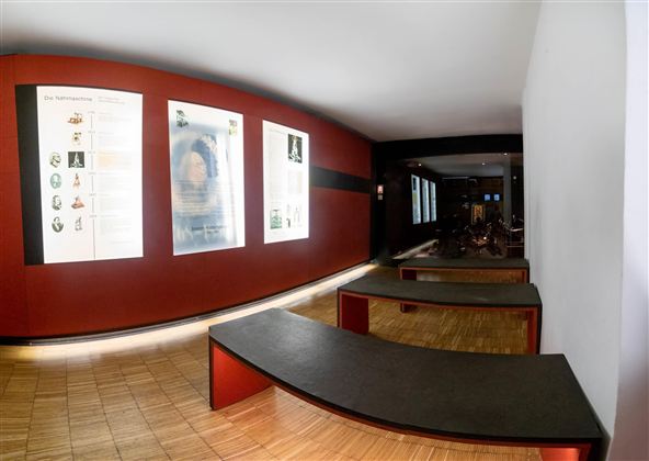 Ein moderner Innenraum mit roten Wänden und Ausstellungsplakaten. Flache Bänke laden zum Verweilen ein.
