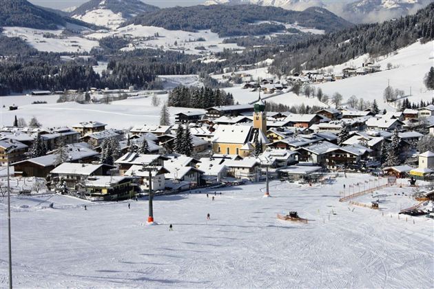 A winter landscape with a small, snowy village and snow-covered mountains in the background. There are skiers on the slope and a beautiful, clear sky.