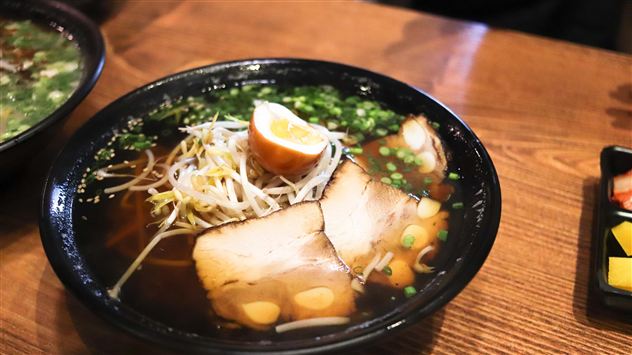 A bowl of ramen with broth, spring onions, and two slices of pork. A piece of lemon and mung bean sprouts are also included.