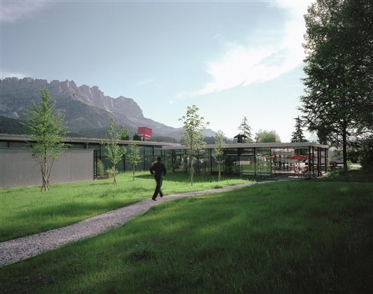 Ein modernes Gebäude in einer grünen Landschaft mit Bergen im Hintergrund. Ein Mann geht auf einem Weg entlang, der durch das Gras führt.