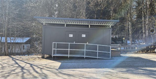 Eine moderne, graue Umkleidekabine steht auf einem befestigten Platz. Umgeben von Bäumen und einem klaren Himmel wirkt sie einladend und funktional.