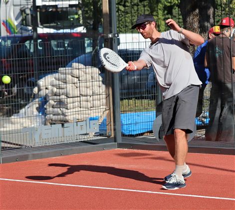 A man is playing padel tennis on a red court. The sun is shining and construction work can be seen in the background.