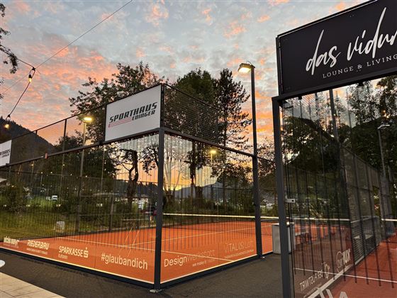 A tennis court with a bright orange surface is surrounded by a fence. In the background, a beautiful sunset and some trees can be seen.