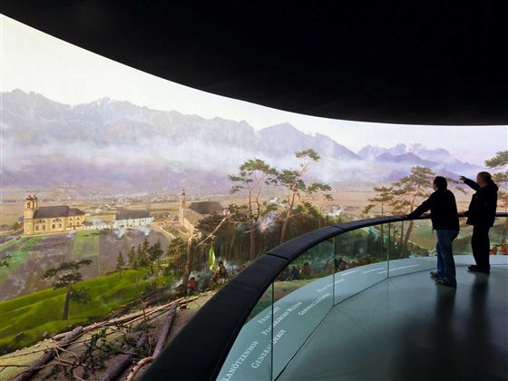 Zwei Personen stehen in einem modernen Ausstellungsraum und schauen auf eine beeindruckende Landschaft mit Bergen und einer kleinen Kirche. Die Aussicht ist lebendig und detailreich, was die Schönheit der Natur hervorhebt.