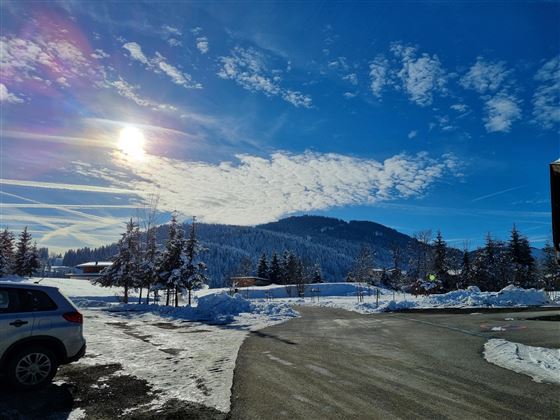 Een besneeuwd landschap met een heldere blauwe lucht en stralende zon. Op de achtergrond zijn beboste bergen te zien.