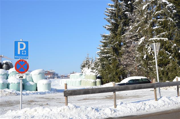 Eine verschneite Landschaft mit einem Parkplatzschild und einem Verbotsschild. Im Hintergrund sind große grüne Ballen und schneebedeckte Bäume zu sehen.