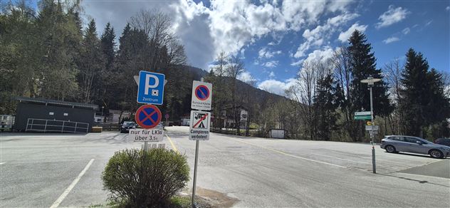 Ein Parkplatz mit verschiedenen Verkehrsschildern in einer grünen Umgebung. Der Himmel ist teilweise bewölkt und der Parkplatz scheint ruhig zu sein.