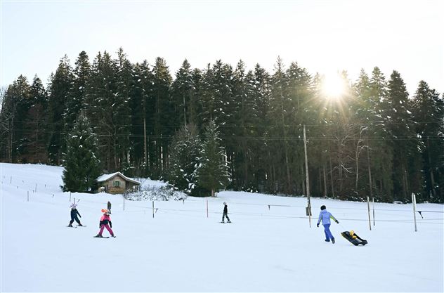 A winter landscape with snow and trees in the background.  
Children are pulling sleds on a frozen area.