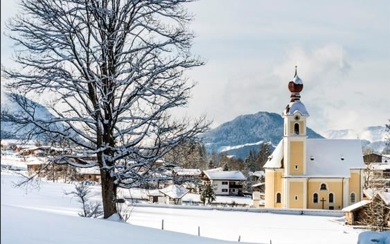 Een schilderachtig sneeuwlandschap met een gele kerk en een grote boom. De bergen op de achtergrond maken het winterse beeld compleet.