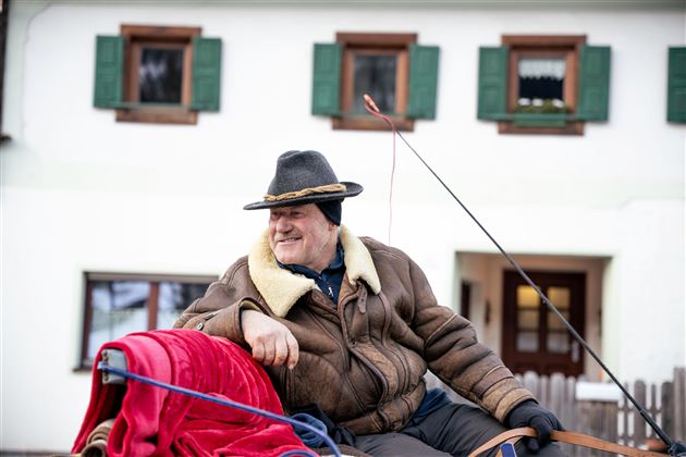 Een glimlachende oudere man zit op een koets. Op de achtergrond zie je een traditioneel huis met groene luiken.