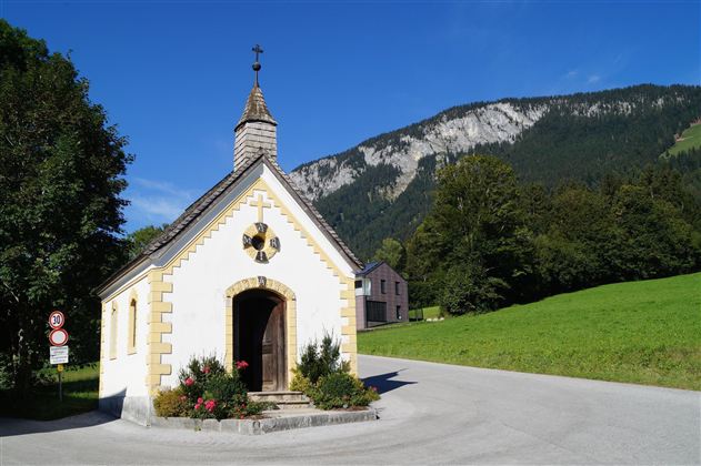 Eine kleine Kapelle steht an einer Straßenecke, umgeben von einer grünen Wiese und Bergen. Der Himmel ist blau und klar.